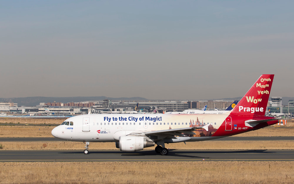 El avión, decorado con el la frase «Fly to the City of Magic!» en el fuselaje y el mensaje «Wow Prague» en la cola rodando por el aeropuerto; al fondo se ven otros aviones y edificios del aeropuerto