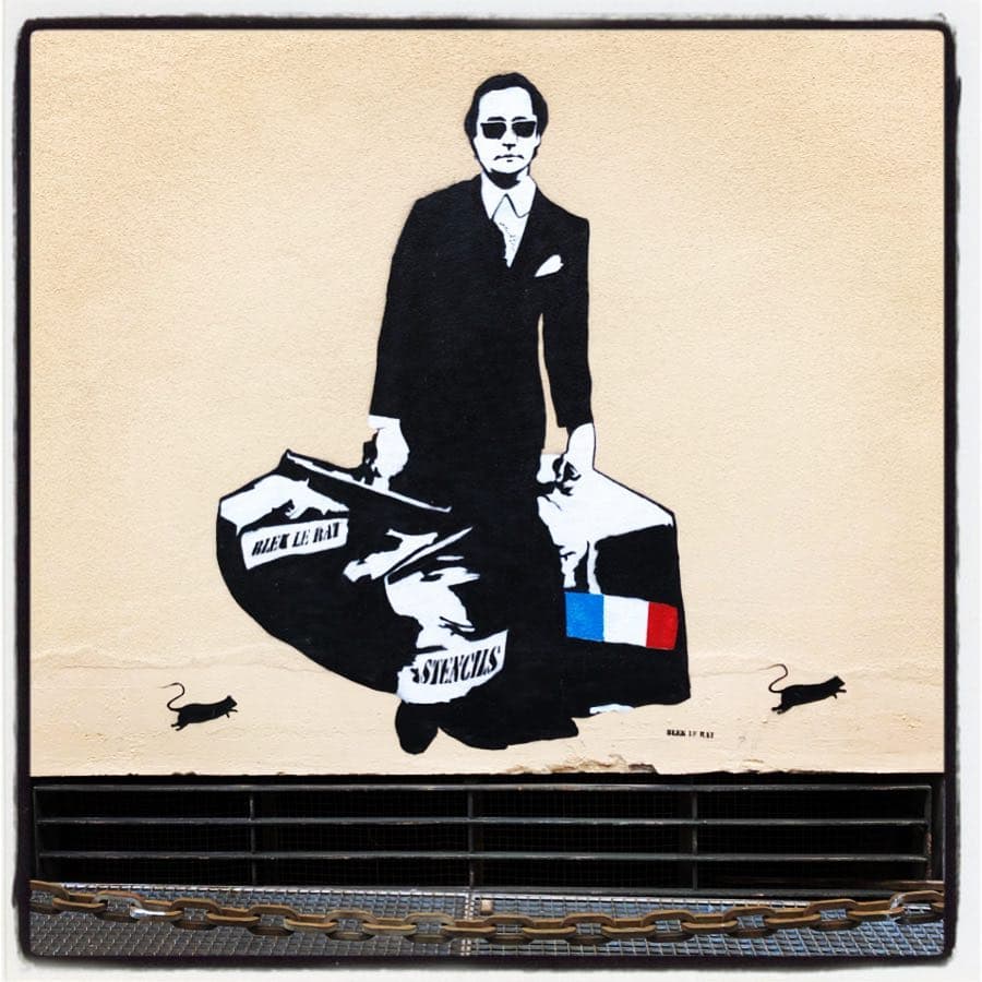 Stencil a tamaño real pegado en una pared pintada de beige en el que un señor trajeado lleva dos maletas etiquetadas con los rótulos «stencils» y «Blek Le Rat» así como una badera francesa; le acompañan un par de las ratas características el autor 